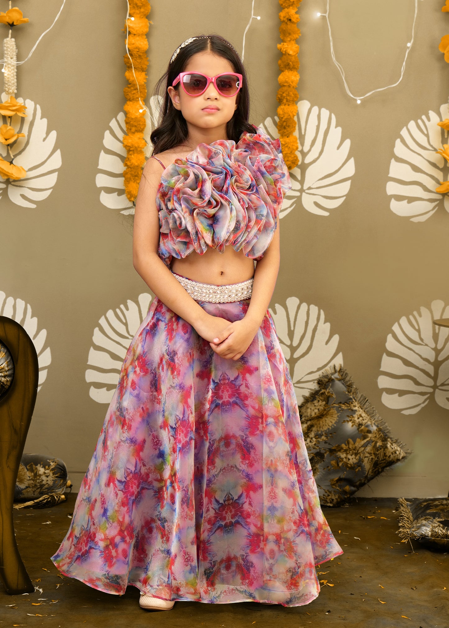 Girl wearing a floral printed lehenga with a statement ruffle top, holding a pose with sunglasses, standing in front of a festive backdrop.