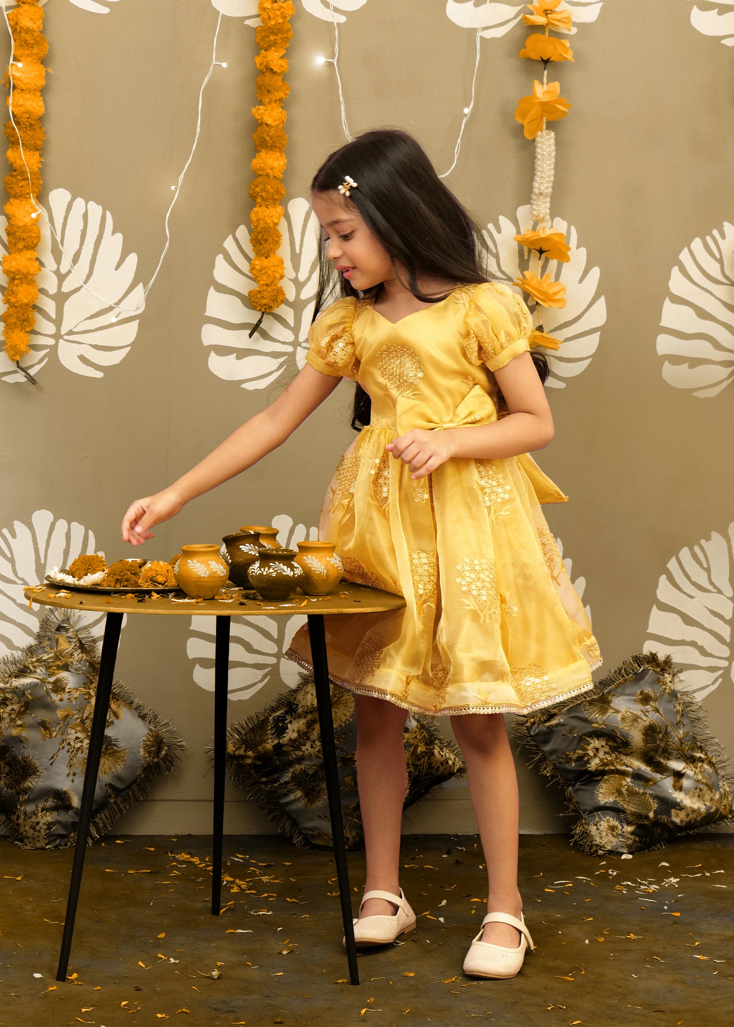 Little girl in a golden embroidered puff-sleeve dress, adjusting festive clay pots on a decorative table.