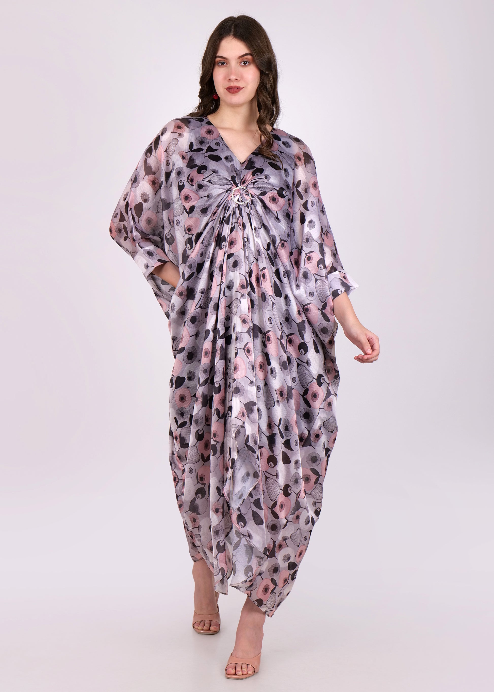 A woman wearing a flowing kaftan dress with a soft floral print in shades of gray, pink, and black. The dress features a V-neckline and a ruched detail at the front.