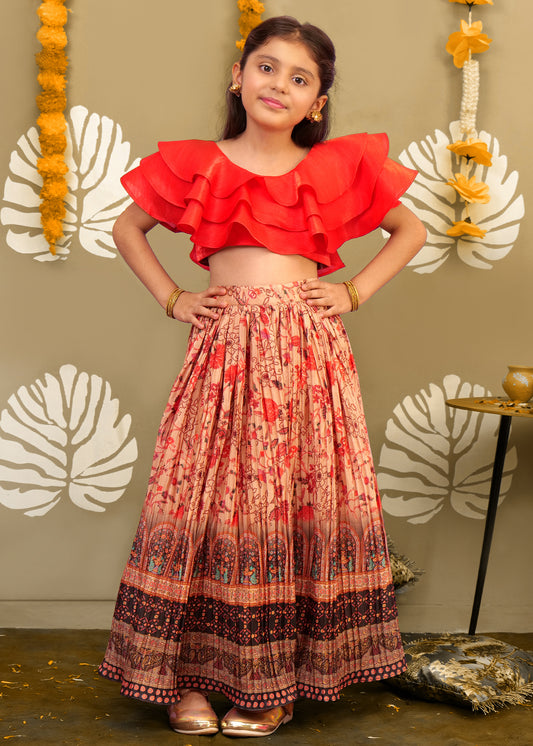 A girl wearing a bold red ruffled top and a printed long skirt in a festive pose, against a traditional decor background.