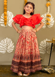 A girl wearing a bold red ruffled top and a printed long skirt in a festive pose, against a traditional decor background.