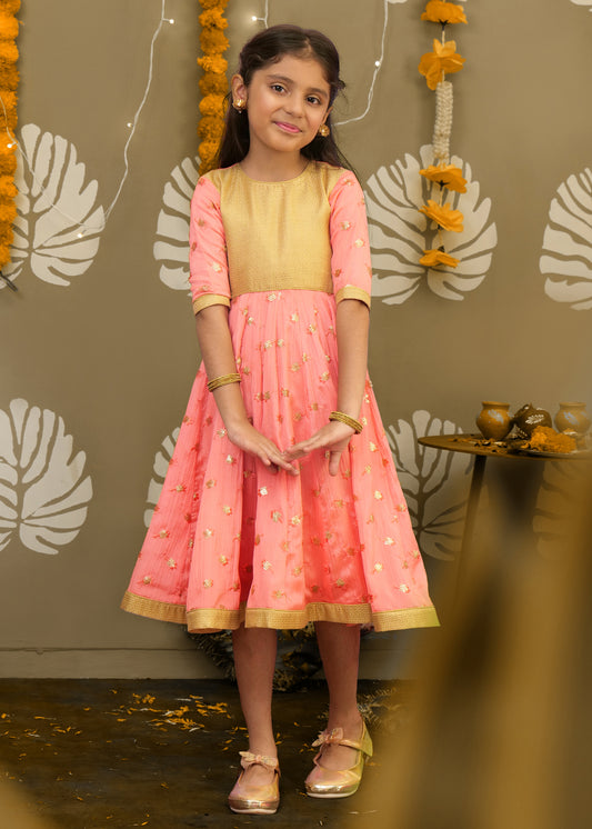 Young girl in a peach and gold embroidered dress posing gracefully in traditional festive attire with golden decor in the background.