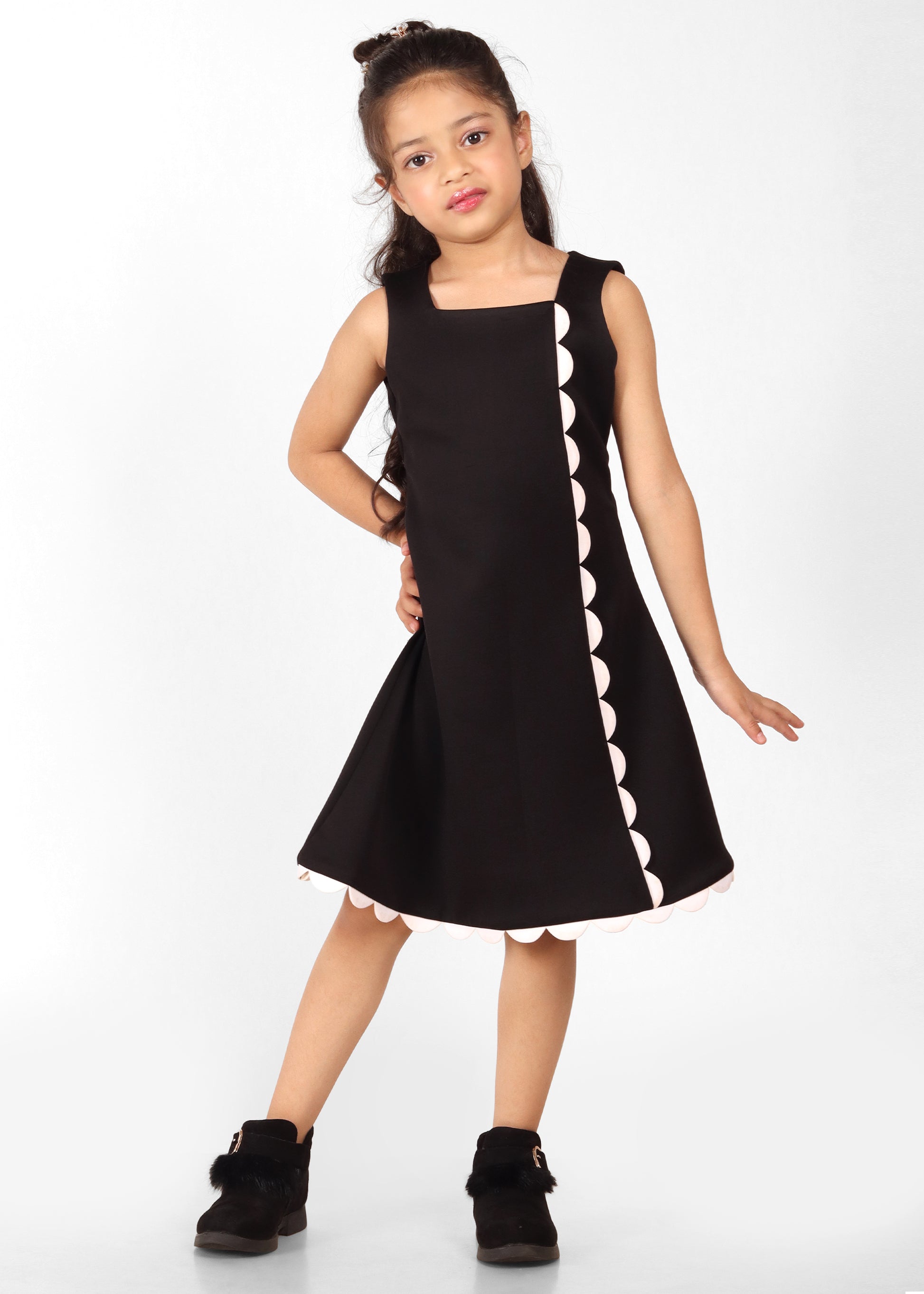 Girl wearing a stylish black sleeveless dress with scallop trim details on the front, standing confidently in black boots