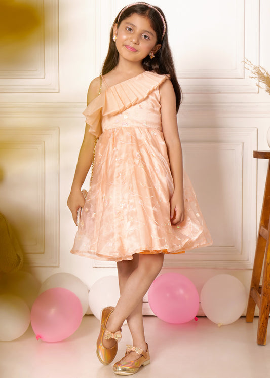 "A young girl wearing a peach-colored one-shoulder dress with pleated ruffles and floral patterns. She is standing against a soft background with pink and white balloons, smiling with one hand on her hip."