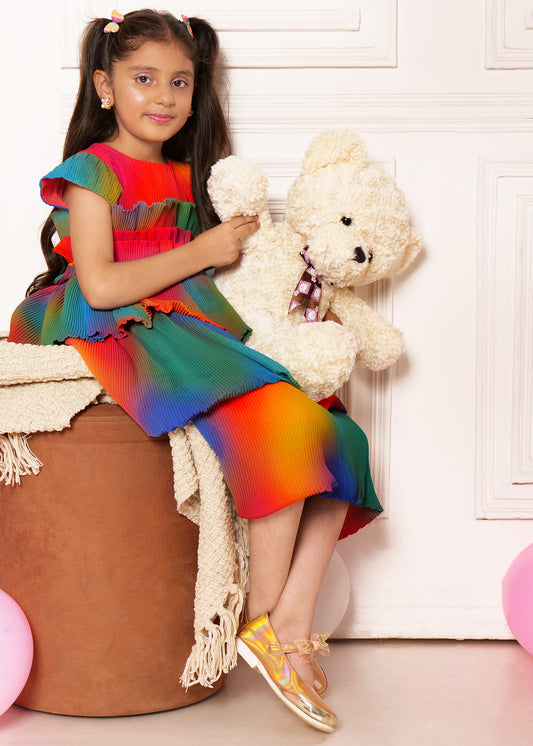 "Girl wearing rainbow pleated dress, holding teddy bear, sitting on a cushioned seat, kids fashion photography."