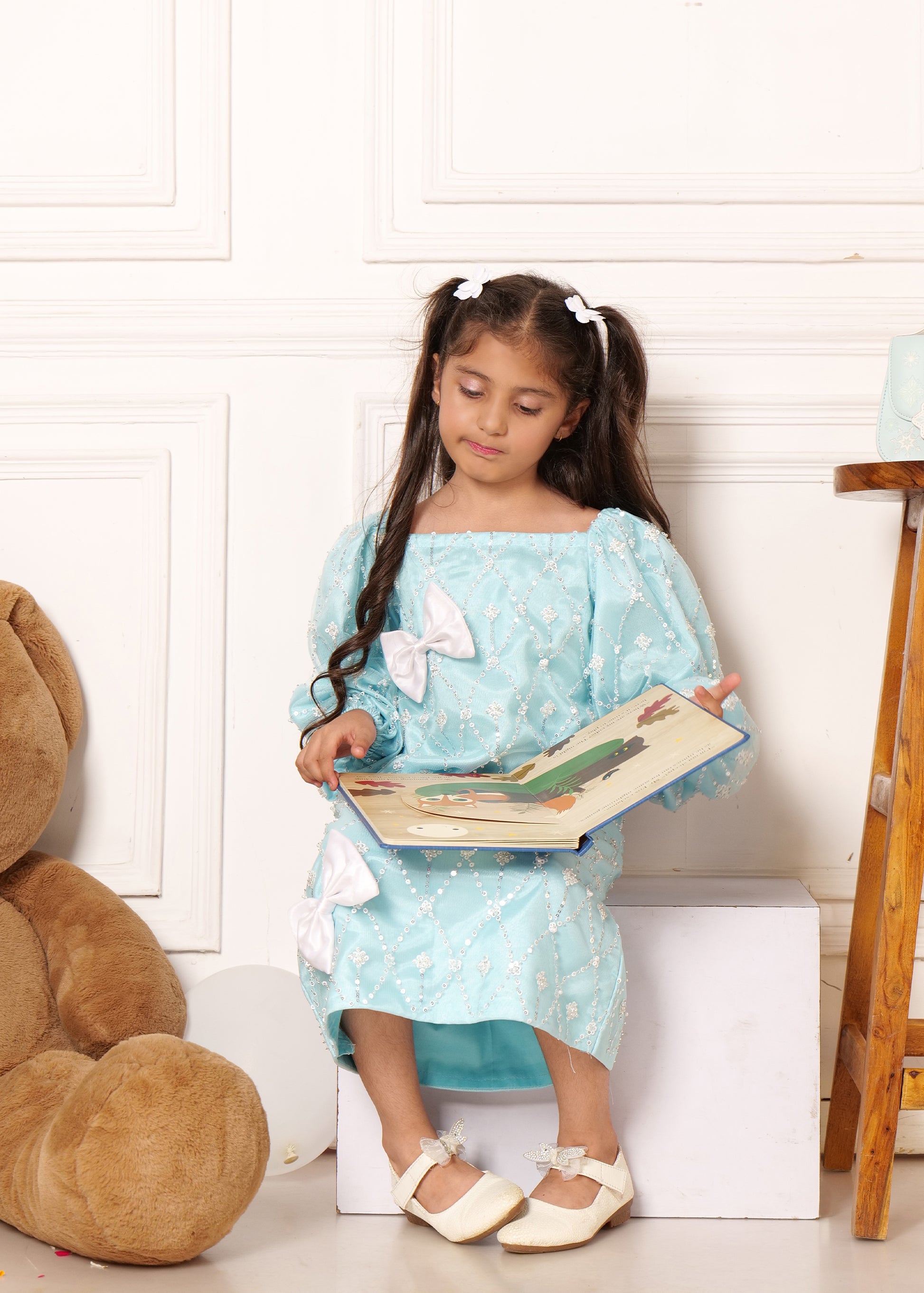  Little girl in a light blue embroidered dress with white bows, reading a book while sitting next to a teddy bear, perfect for children's fashion shoots