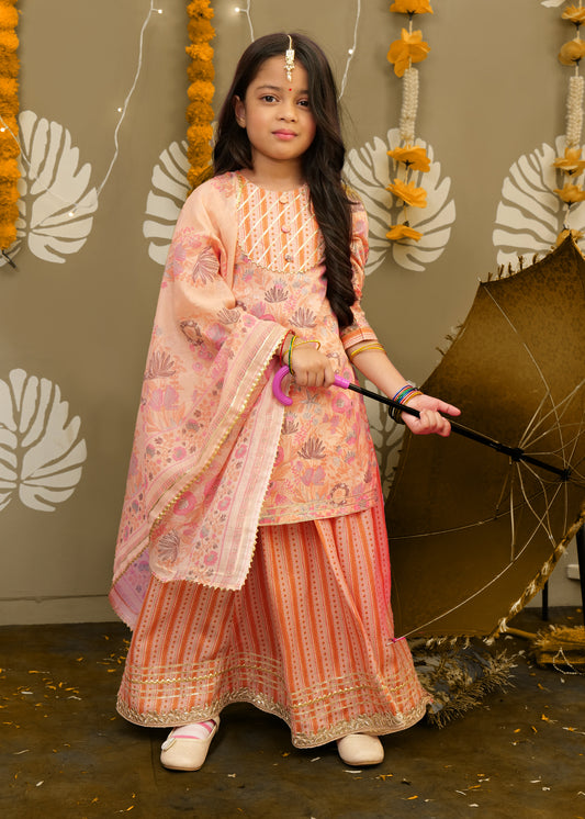 A young girl is wearing a peach-colored floral ethnic outfit featuring a flowy skirt and dupatta, accessorized with bangles, and holding a brown traditional umbrella.