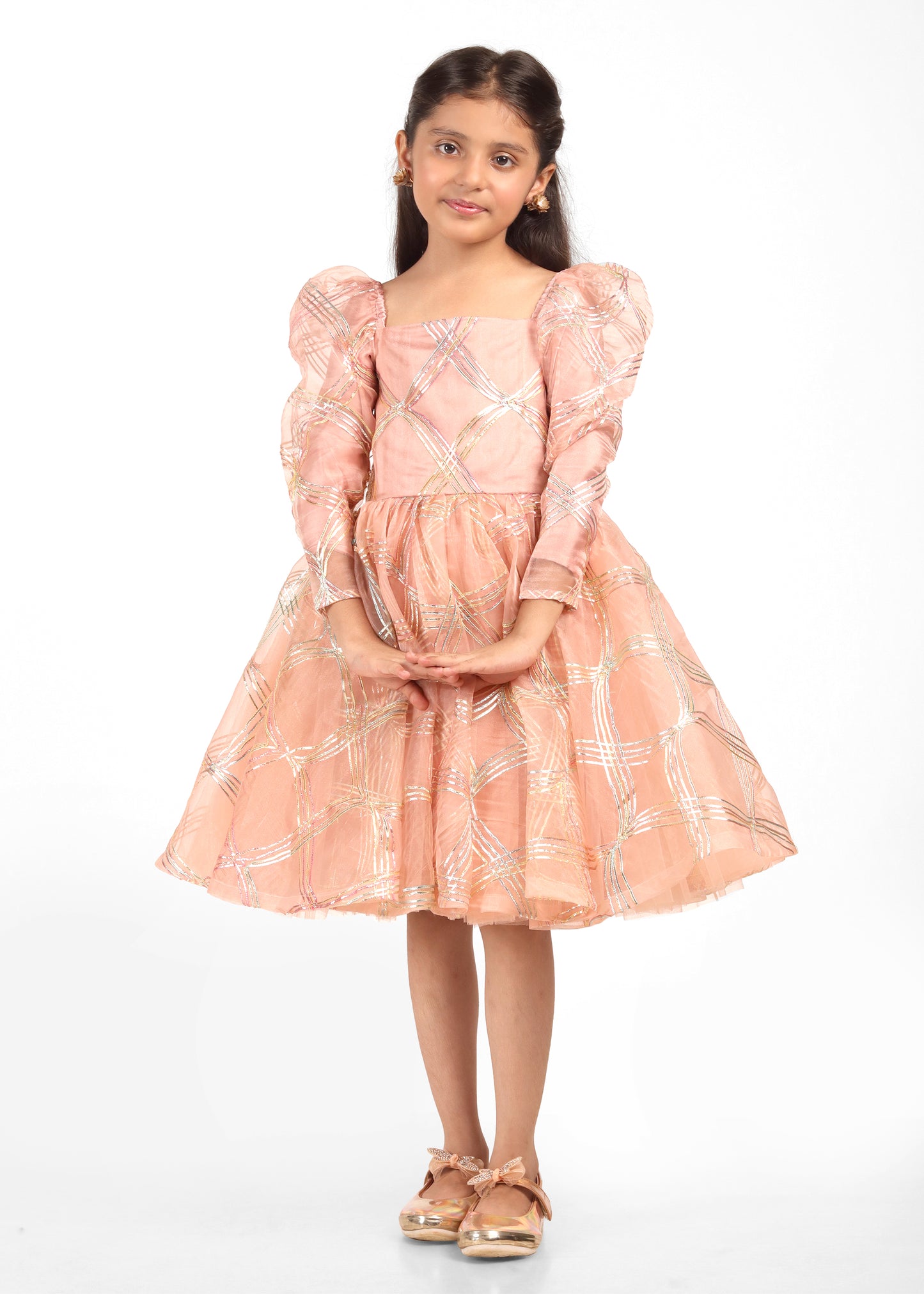 Little girl in a peach and gold puff sleeve dress with intricate golden checkered patterns, posing with elegance and grace