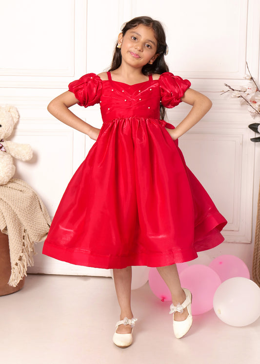 Adorable young girl wearing a vibrant red party dress with puff sleeves and pearl detailing, standing confidently in front of a teddy bear, exuding charm and elegance."