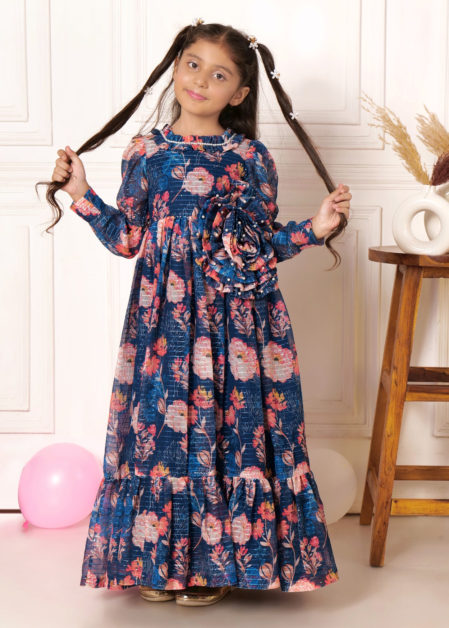 Girl in a blue floral print long dress with puffed sleeves, posing with her hair styled in pigtails, standing near a wooden stool.