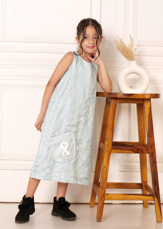 Young girl posing in a light blue sleeveless midi dress with subtle floral embroidery, leaning against a wooden stool, styled with black boots for a chic l