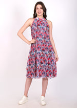 Model wearing a sleeveless, pleated, high-neck printed midi dress in vibrant red and blue patterns, paired with white sneakers.