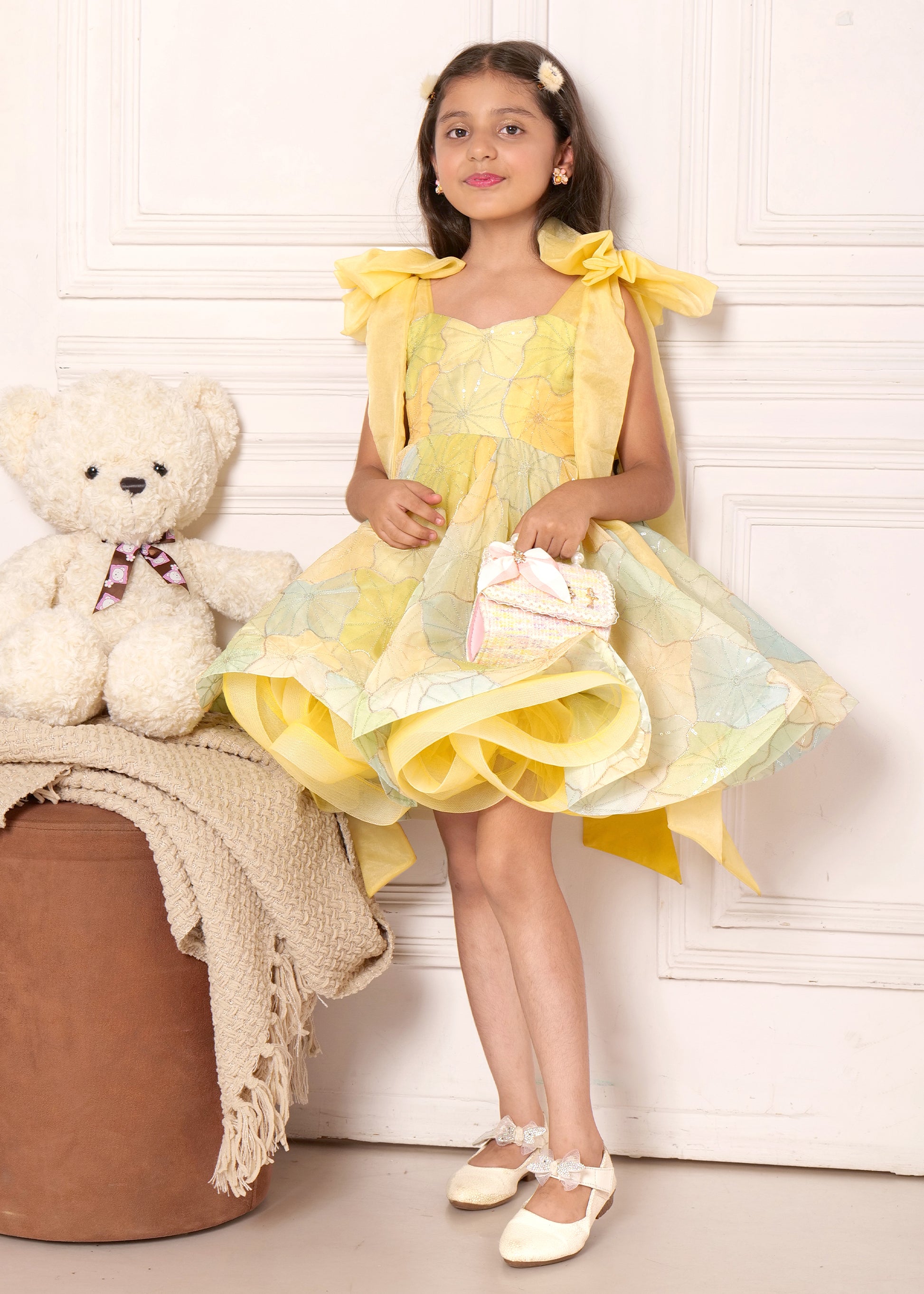 "Charming girl wearing a pastel yellow floral dress with large bow straps, standing next to a teddy bear and holding a small purse, exuding elegance."