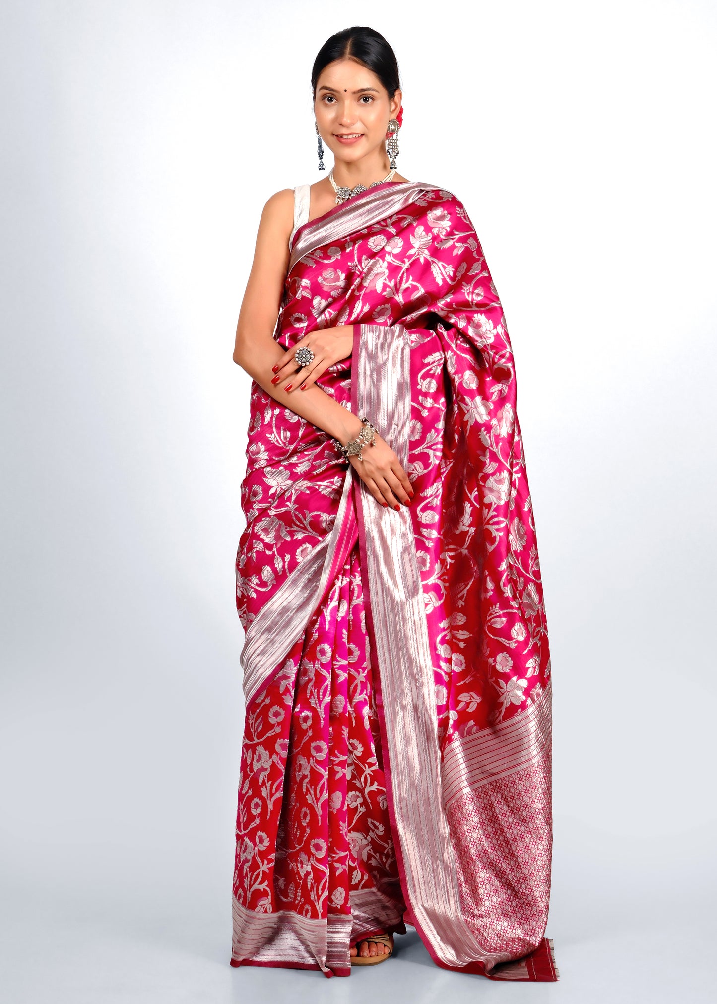 A model wearing a vibrant pink and silver Banarasi silk saree with intricate floral motifs and a shimmering border, styled with traditional silver jewelry.