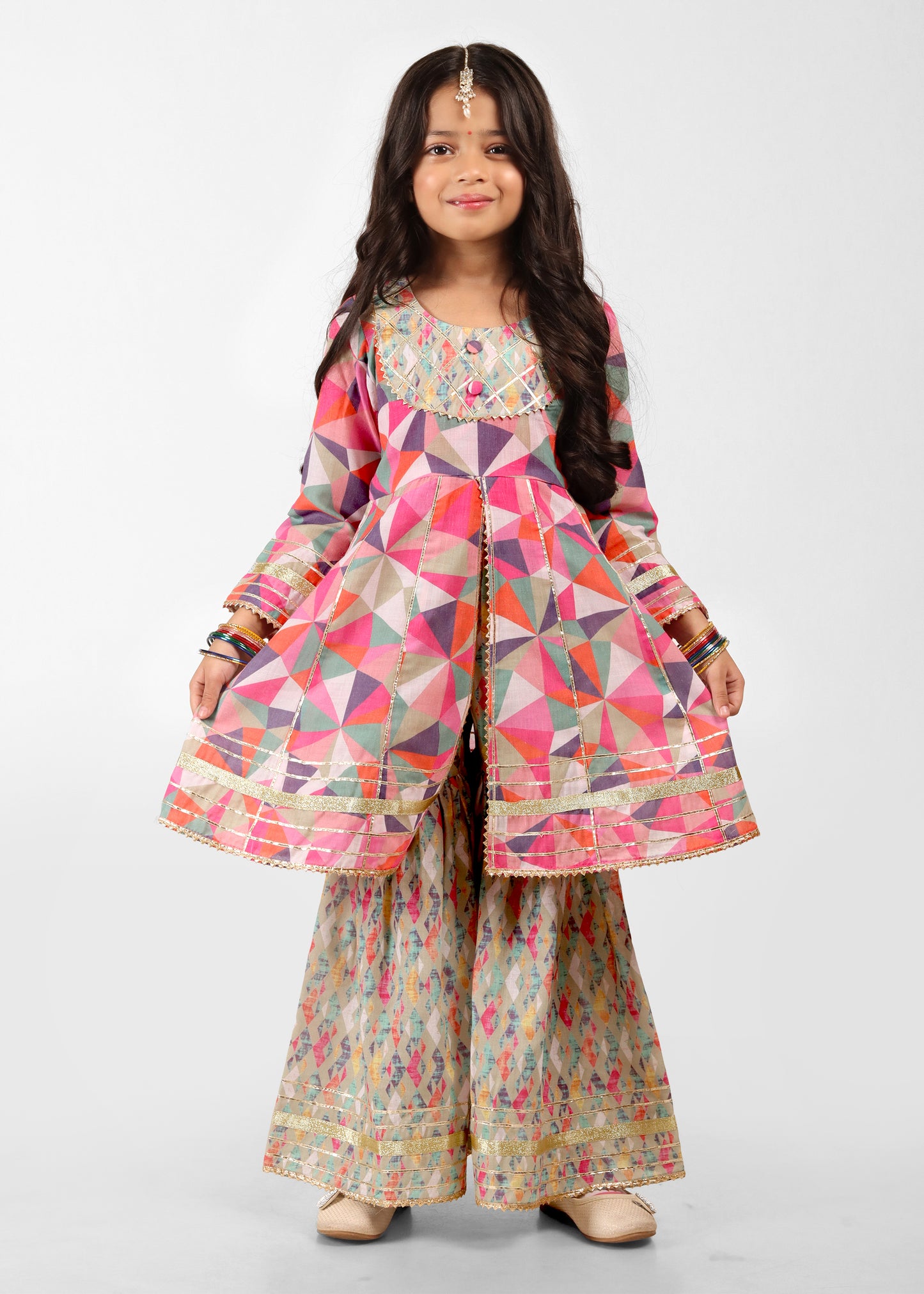 A young girl dressed in a vibrant geometric patterned kurta paired with a matching floral palazzo and dupatta, perfect for festive occasions. The outfit features gold accents and bright colors, creating a playful, traditional look for kids.