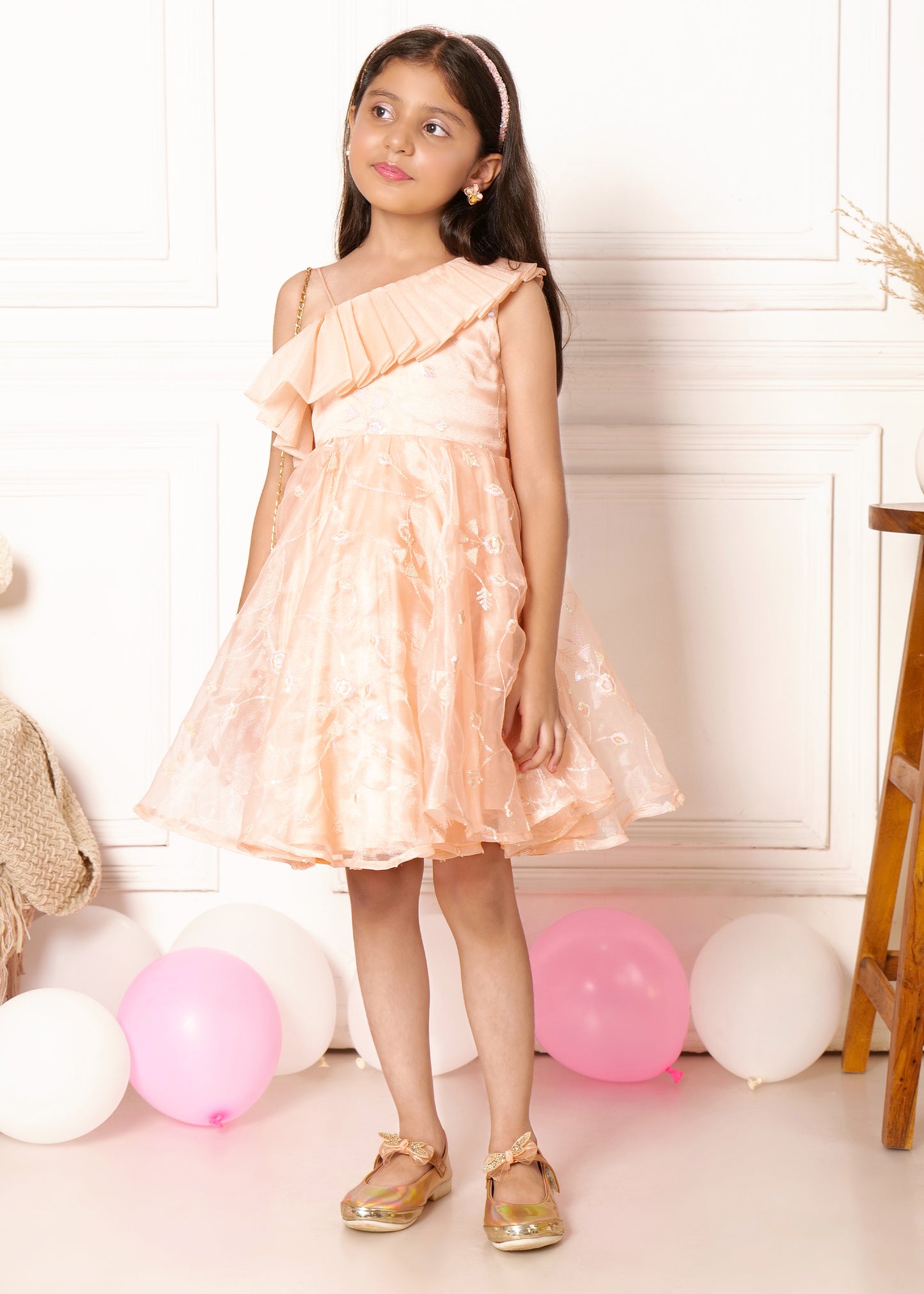 "A young girl dressed in a peach one-shoulder dress adorned with floral details and ruffled neckline, posing next to pink and white balloons while leaning on a chair with a soft smile."