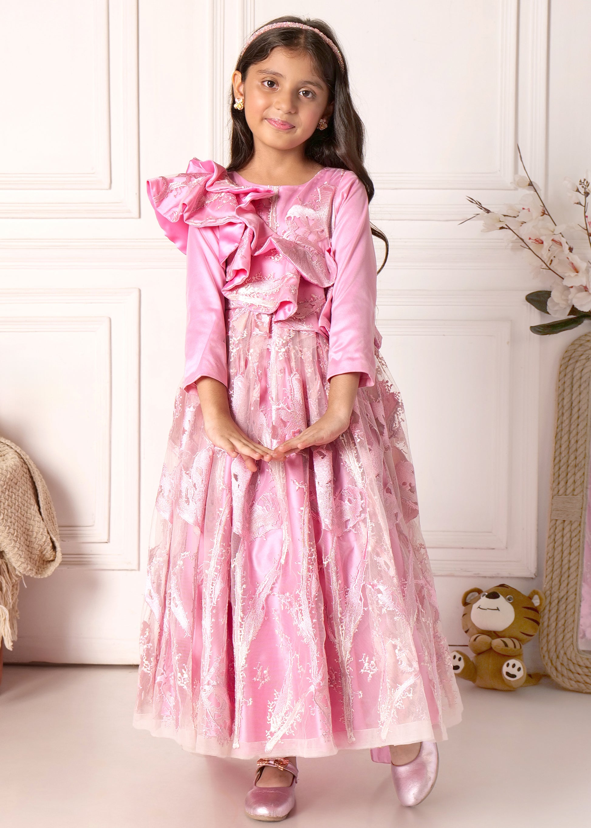 A girl in a pink full-length gown featuring layered ruffles and floral lace, posing with a soft smile.