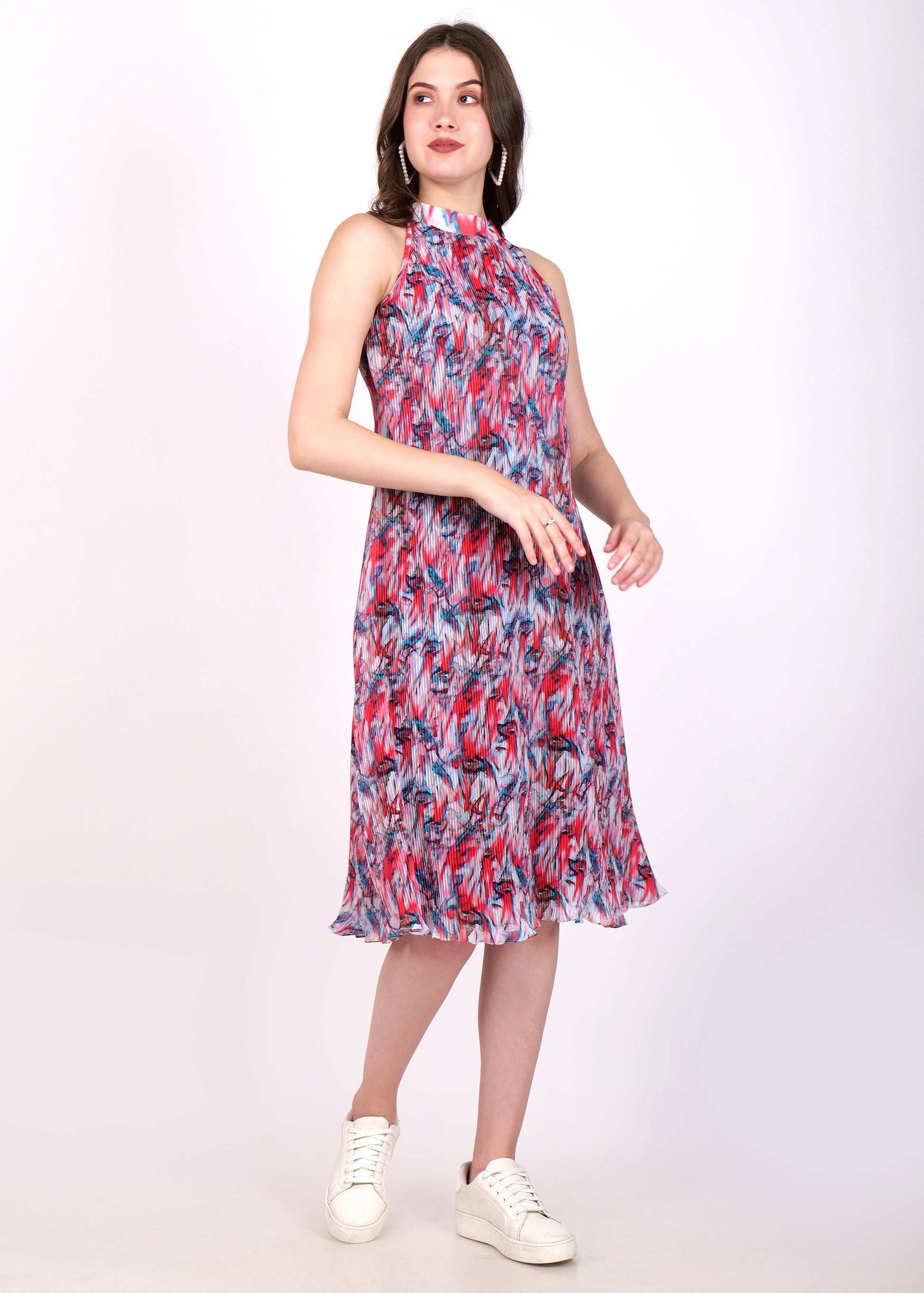 Model posing in a sleeveless, high-neck pleated midi dress with a flowing red and blue print, styled with casual white sneakers.