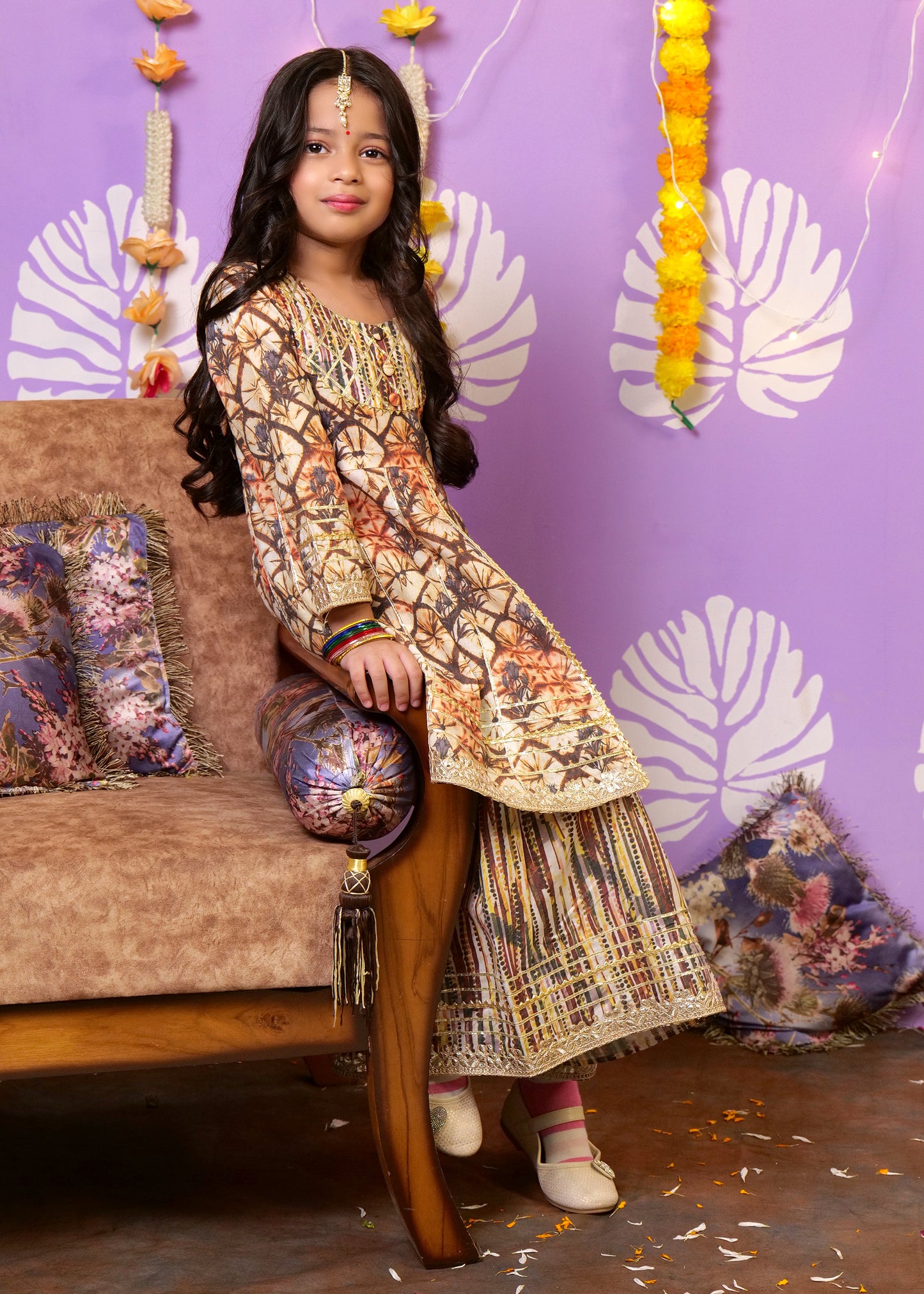 Girl sitting on a couch, dressed in a colorful Indian ethnic dress with marigold garlands in the background, enjoying a festive moment.