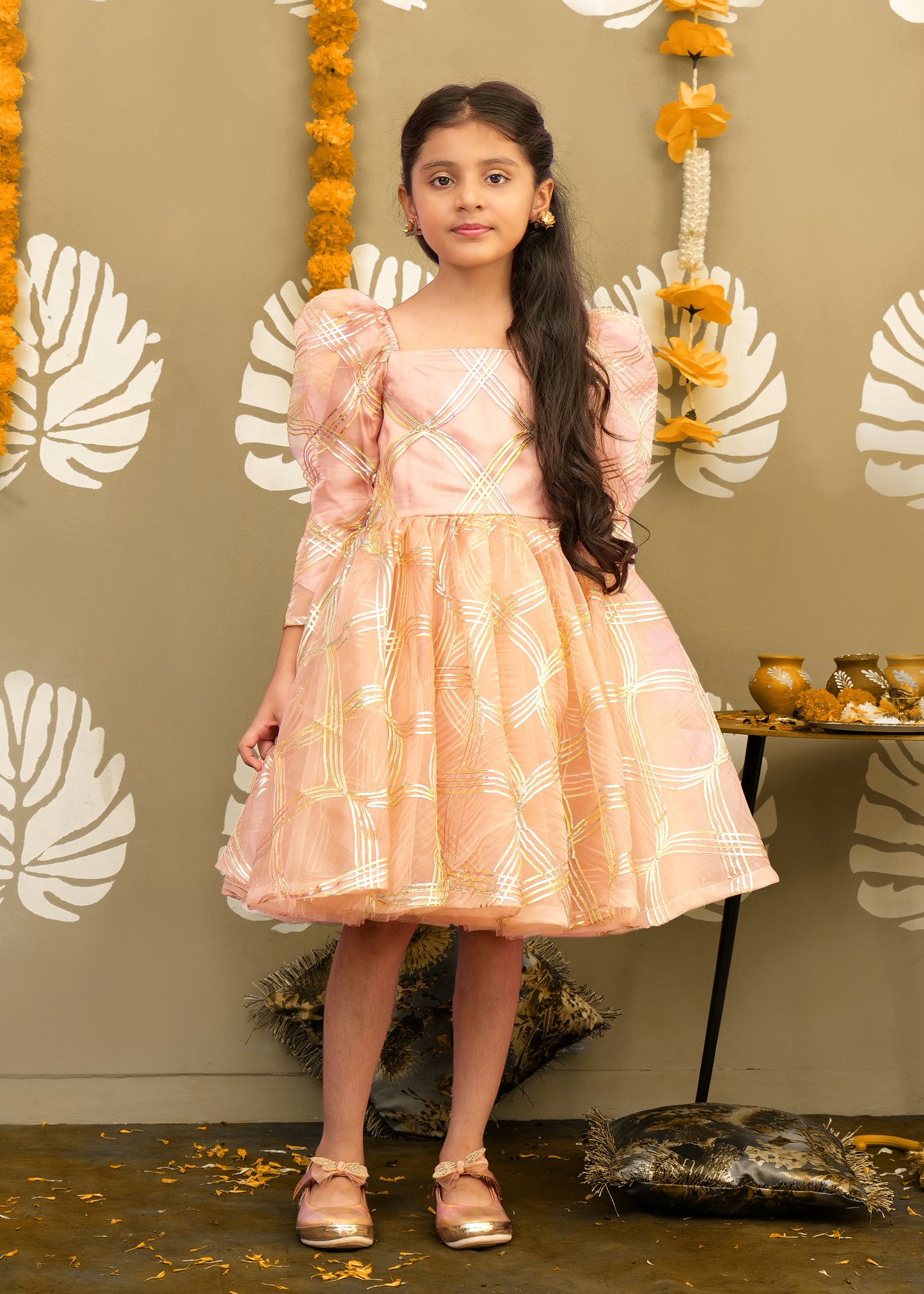 Young girl wearing a peach puff sleeve dress with golden checkered details, standing confidently in front of traditional festive decorations.