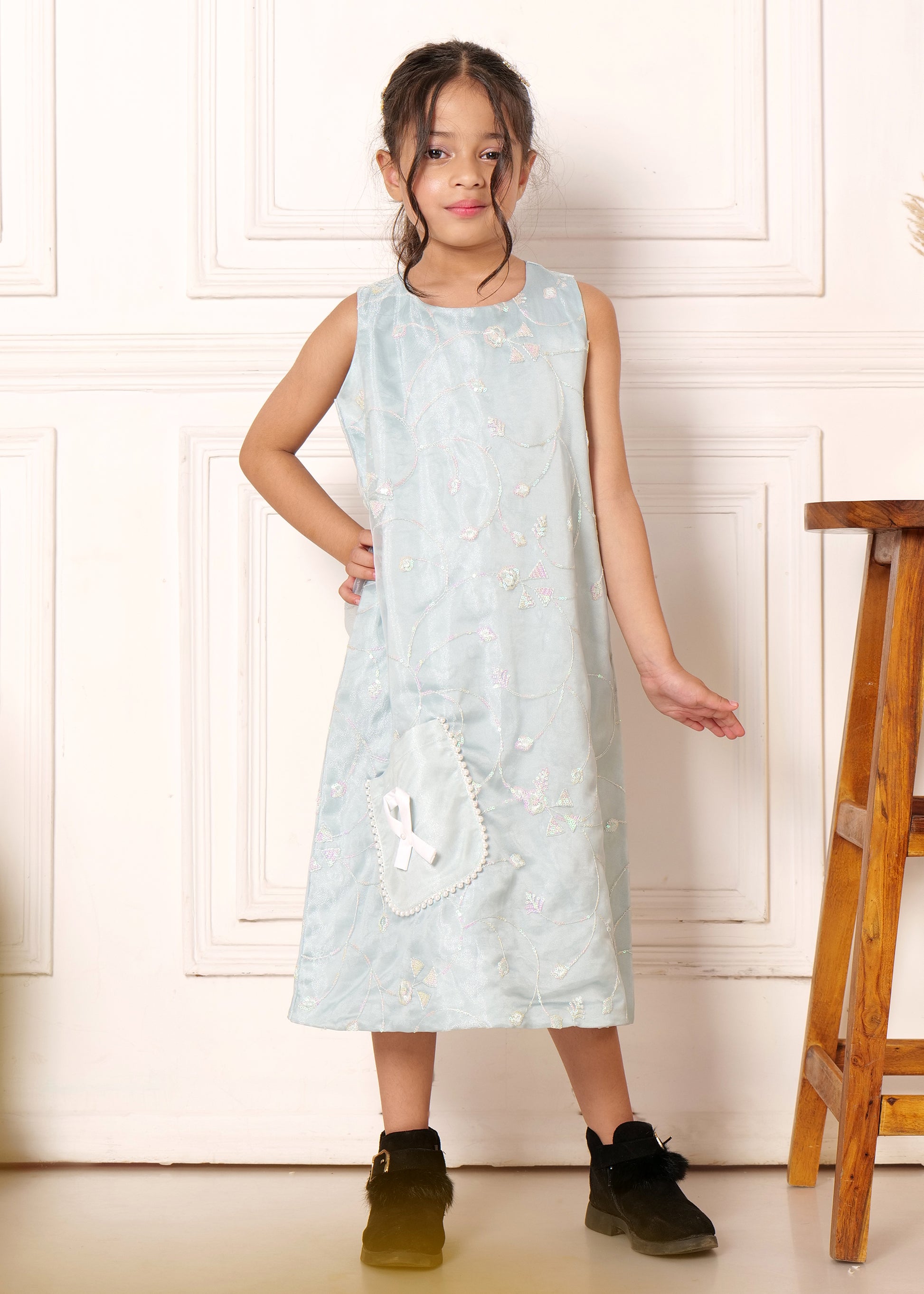 Girl showcasing a sleeveless light blue midi dress with delicate embroidery and white ribbon pocket detail, styled with black ankle boots, exuding a casual elegance