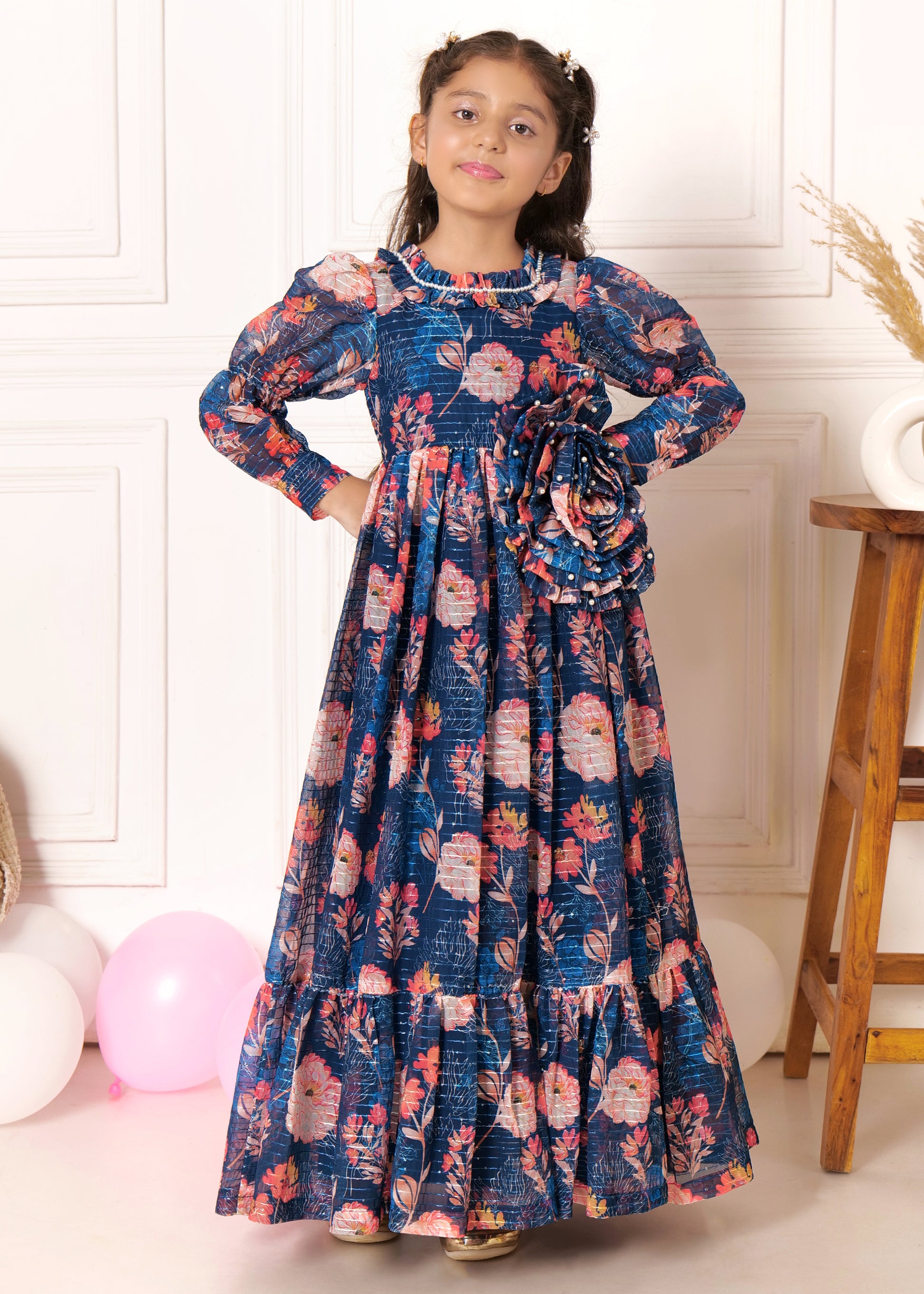 Little girl wearing a blue floral print long dress with puffed sleeves, posing with pigtail hairstyle, standing
beside a wooden stool, perfect for kids' fashion photoshoot or floral dress collection for girls.