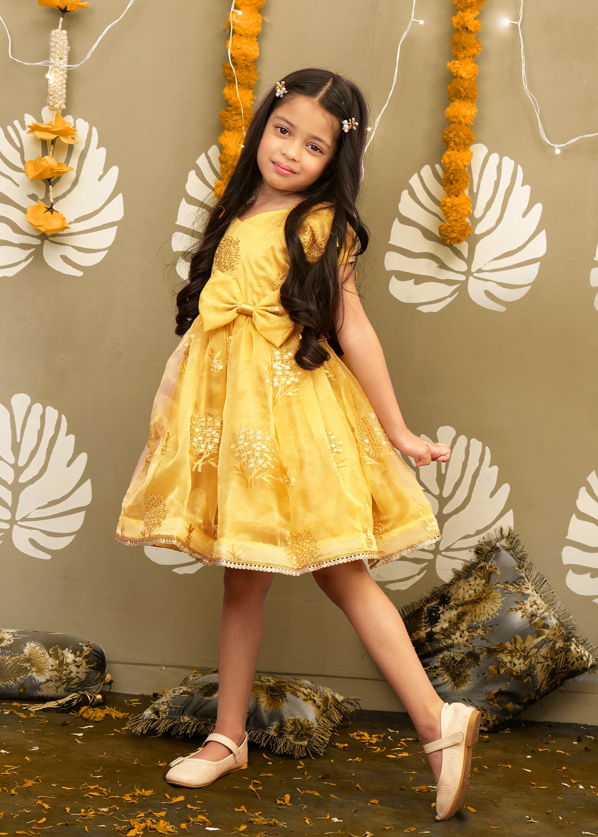 A young girl in a golden festive dress posing gracefully in a traditional decor setting with floral garlands.