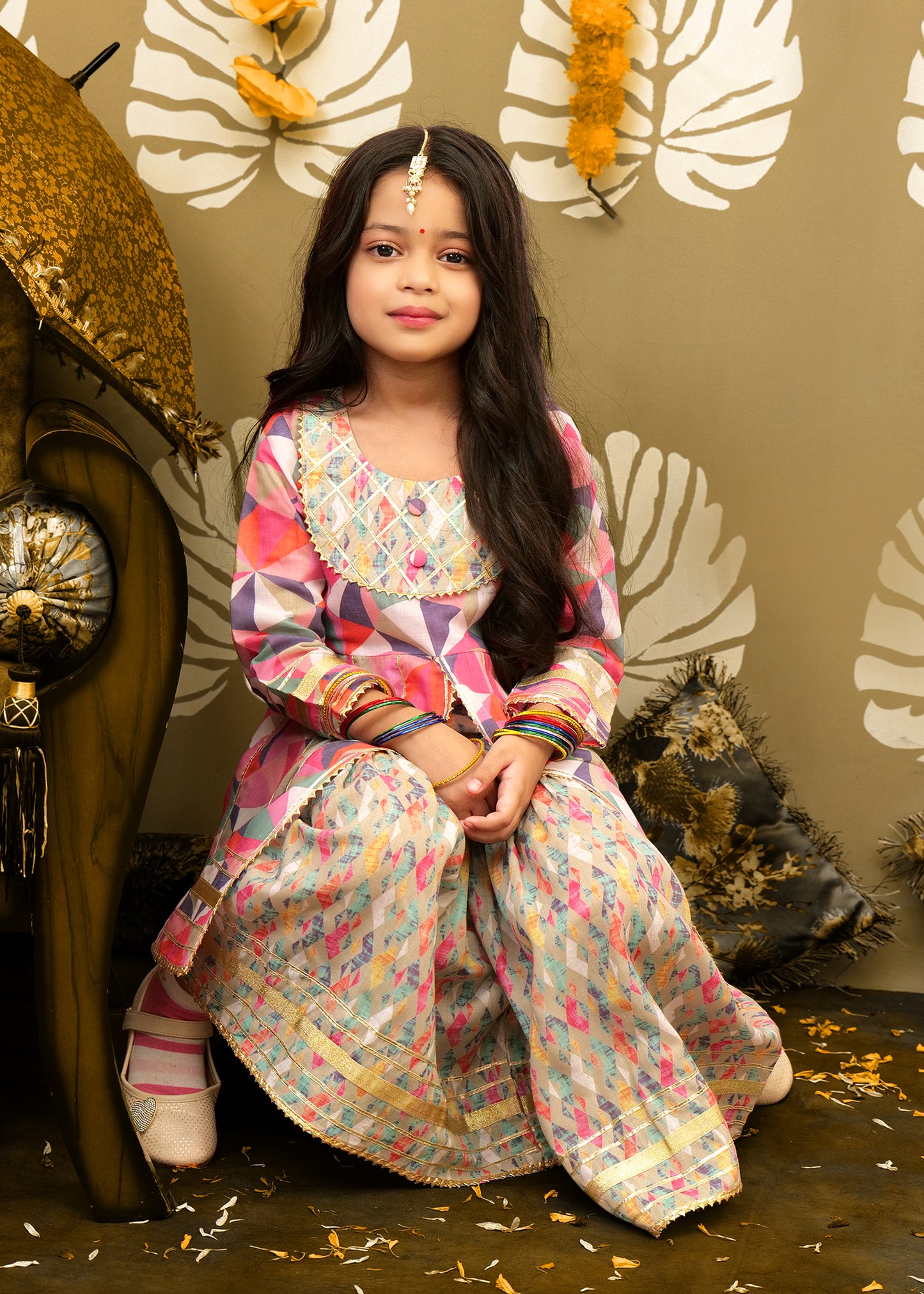 A young girl sitting in a traditional festive setting, wearing a vibrant geometric print kurta and floral palazzo. The ethnic outfit is highlighted by intricate embroidery, suitable for Indian festivals.