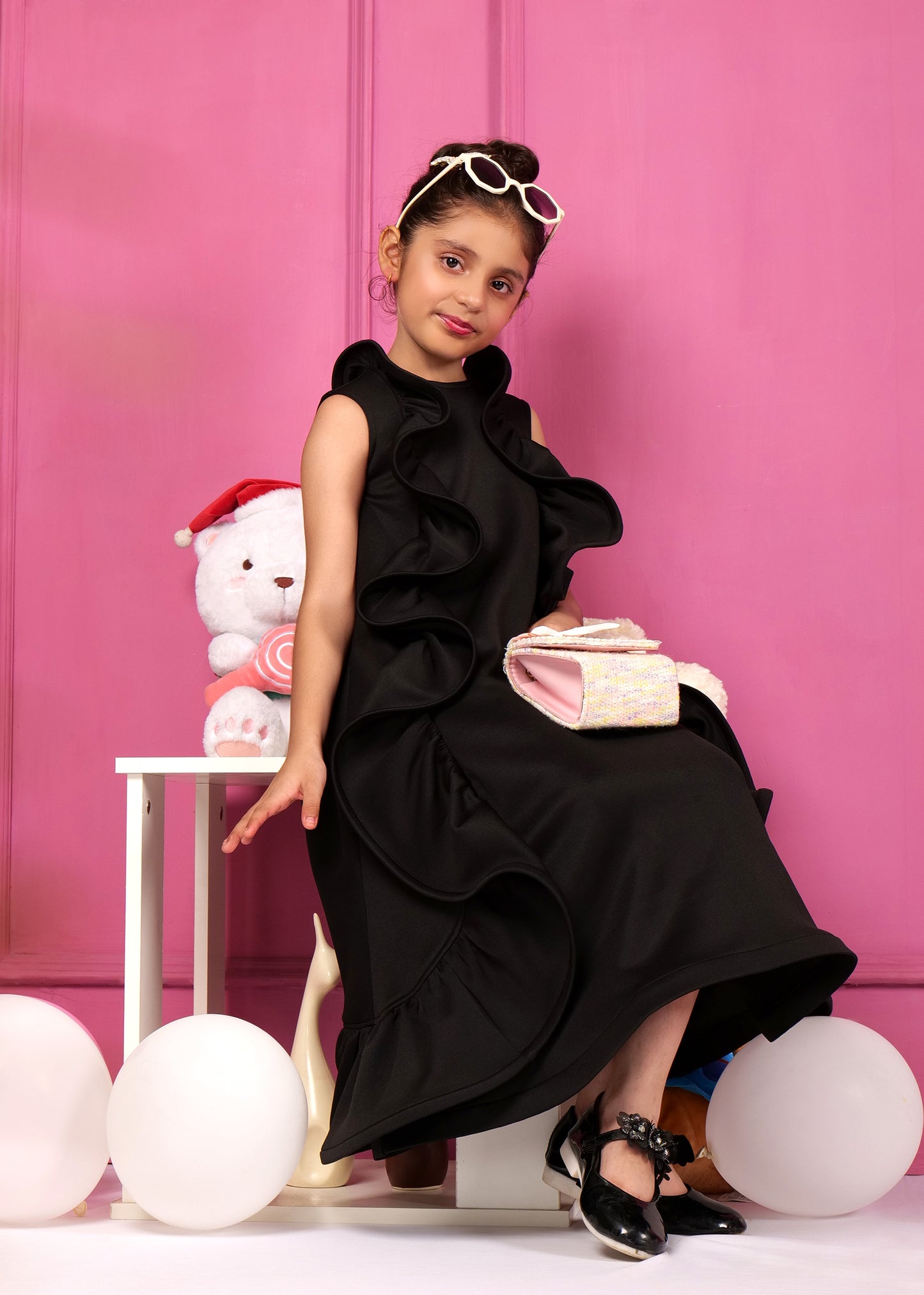 Fashionable girl sitting on a chair in a black sleeveless ruffled dress with sunglasses, holding a chic bag and surrounded by toys