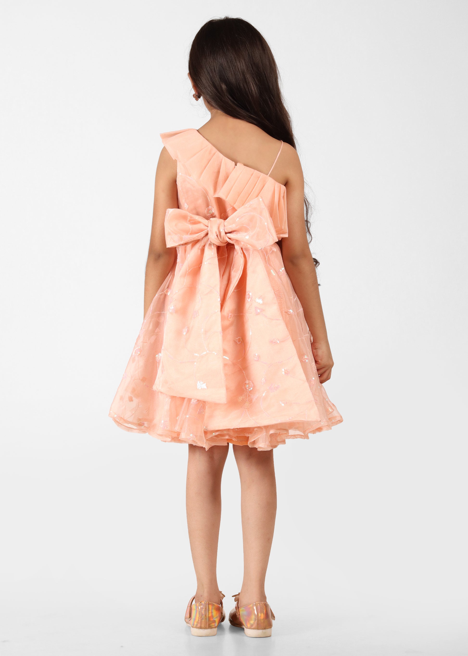 "Back view of a young girl wearing a peach one-shoulder dress with a large bow at the waist, showing off the flowing design of the dress."