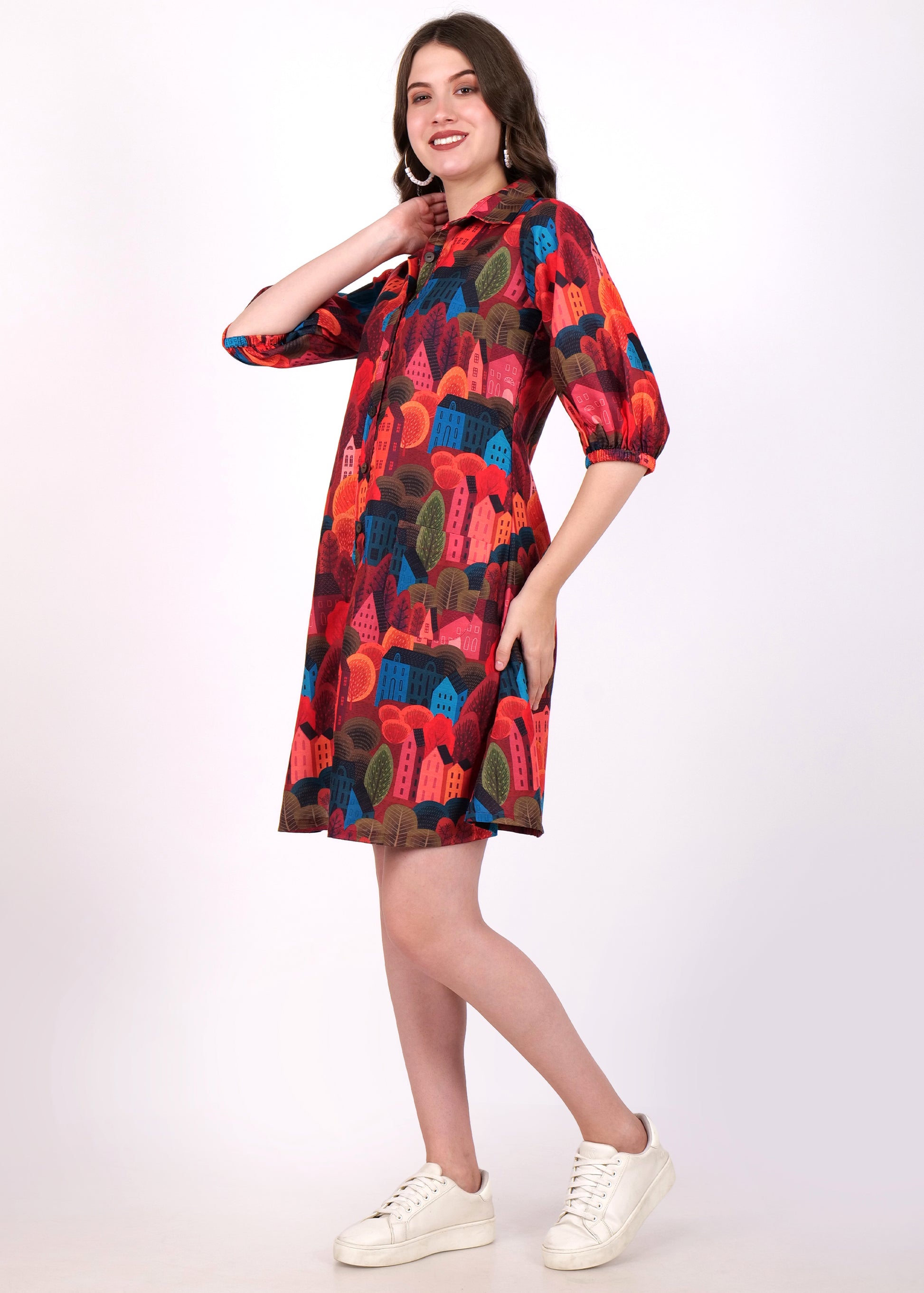  "Side view of a model in a colorful abstract house print shirt dress with vibrant red and blue tones, paired with casual white sneakers."