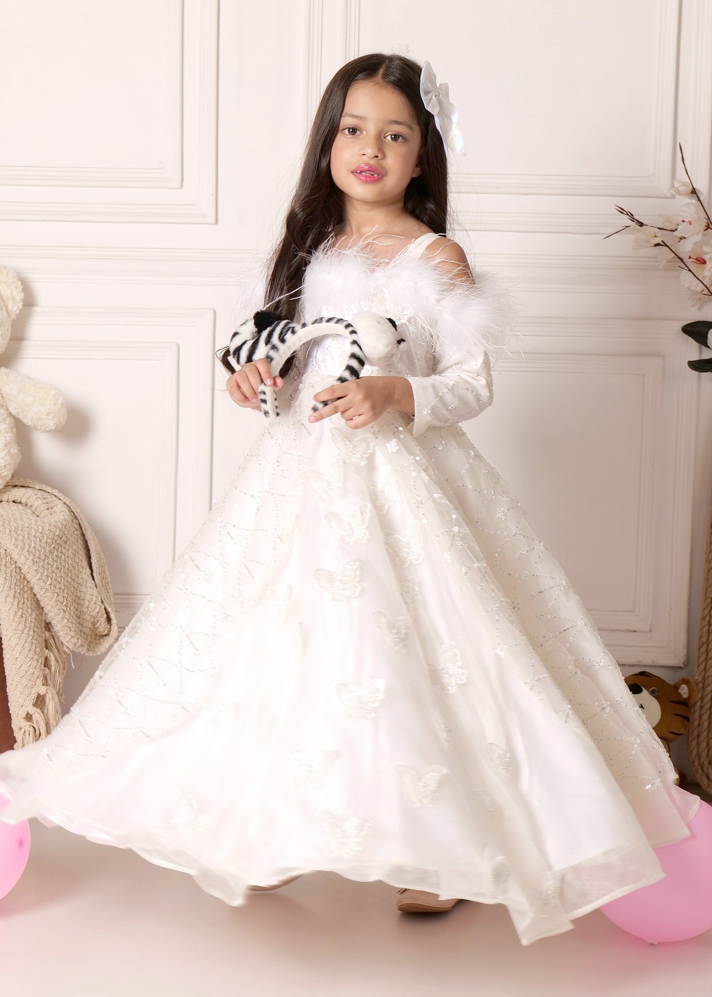 Little girl wearing a white butterfly-sequined gown with long sleeves and feather detailing, standing elegantly in a white room.