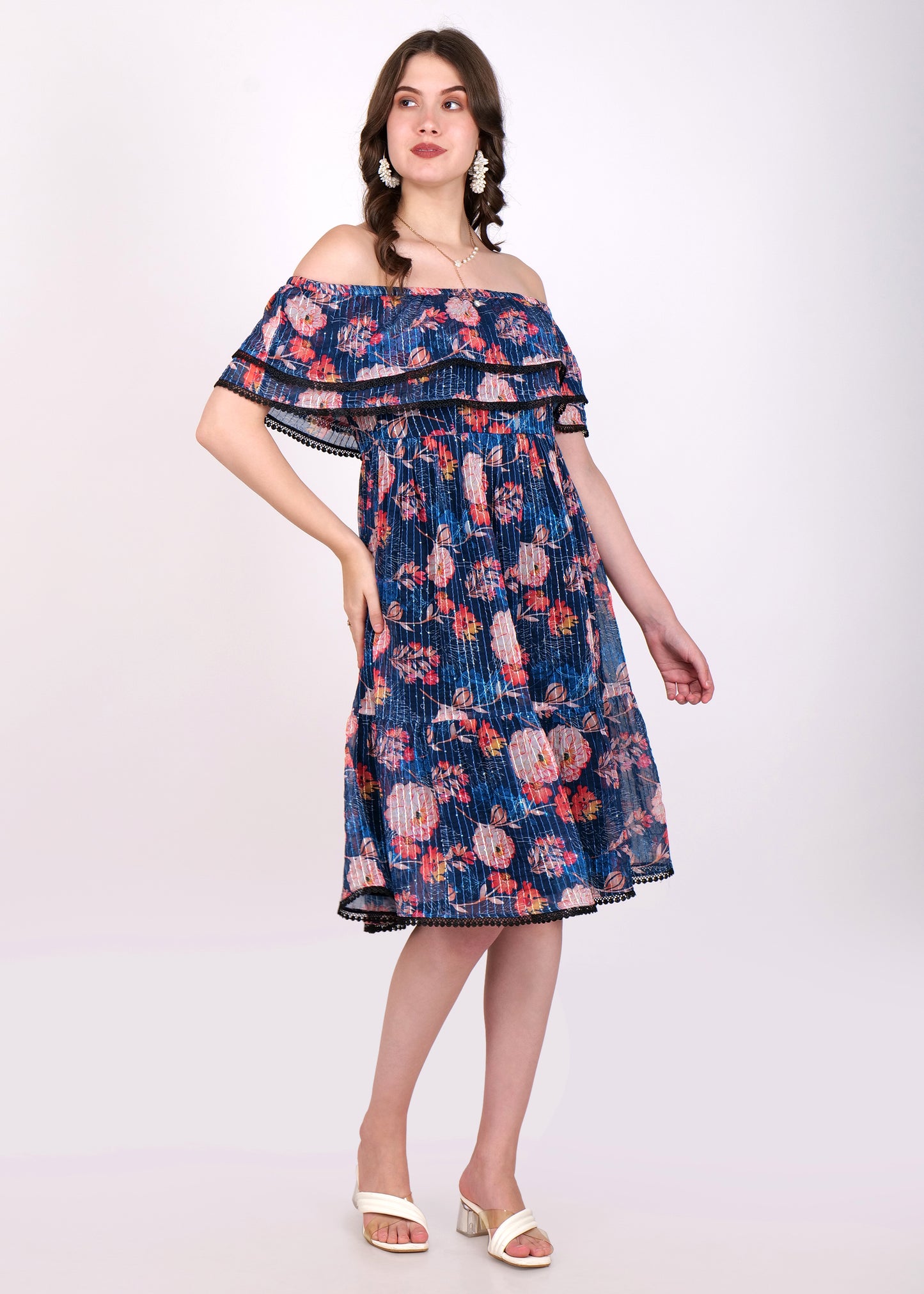 Front view of a woman in a navy blue floral off-shoulder pleated dress featuring tiered ruffles and black lace detailing, paired with white heels