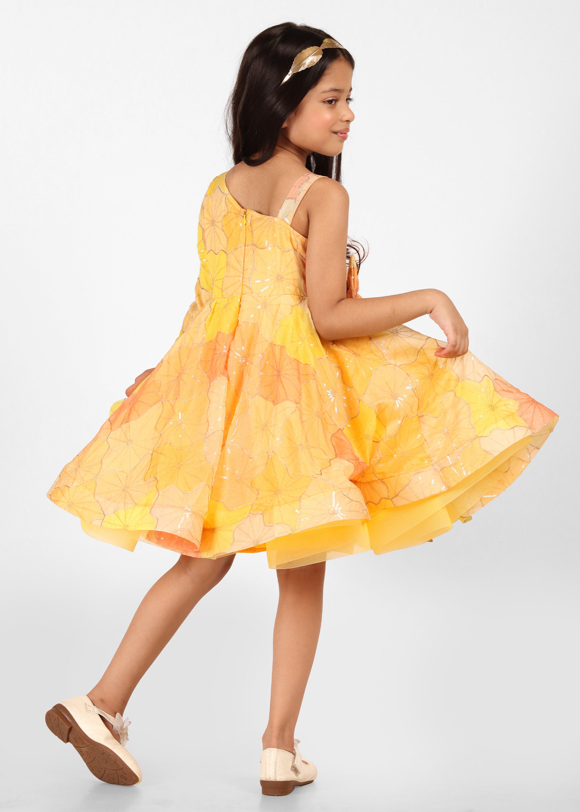 "Charming back view of a young girl twirling in a yellow floral dress with an elegant bow and layered hem."