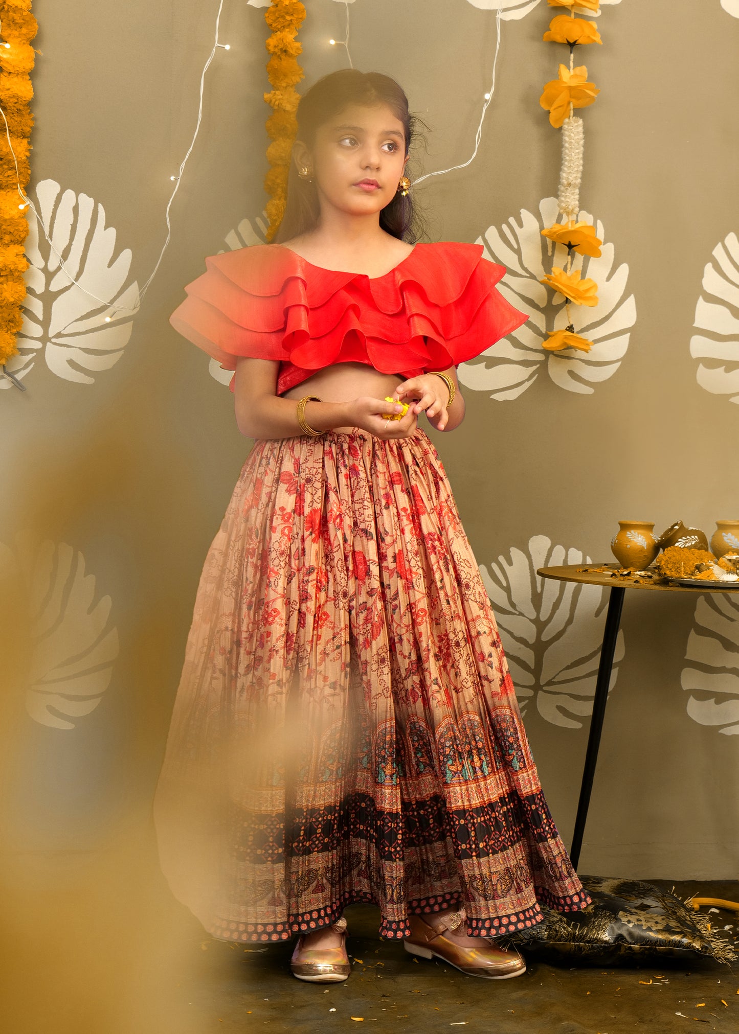 The girl standing gracefully in her printed long skirt and a ruffled red top, adding a modern flair to traditional attire with marigold flower decorations in the background.