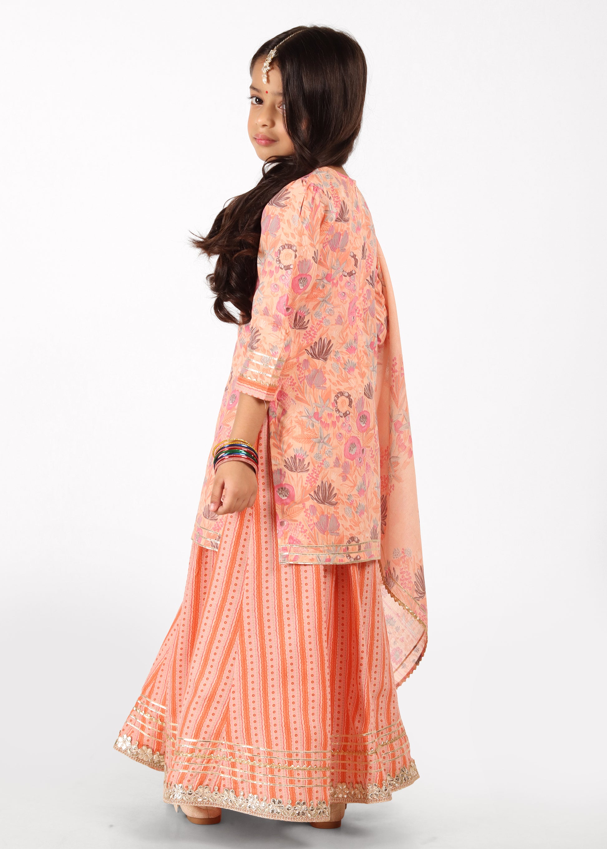 Back view of a girl dressed in a peach lehenga set with floral prints, showing the intricate detailing of the fabric and flowy silhouette.