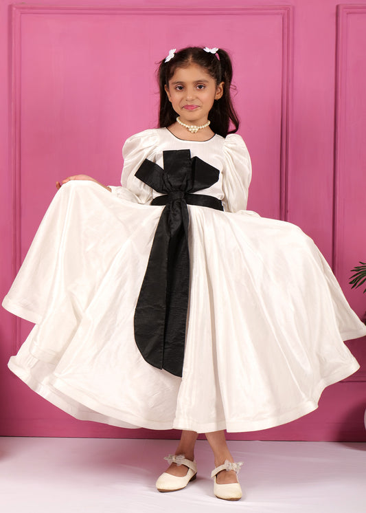 Cute girl displaying a white puff-sleeve dress with a black bow, holding the dress wide as she smiles in front of a pink backdrop.