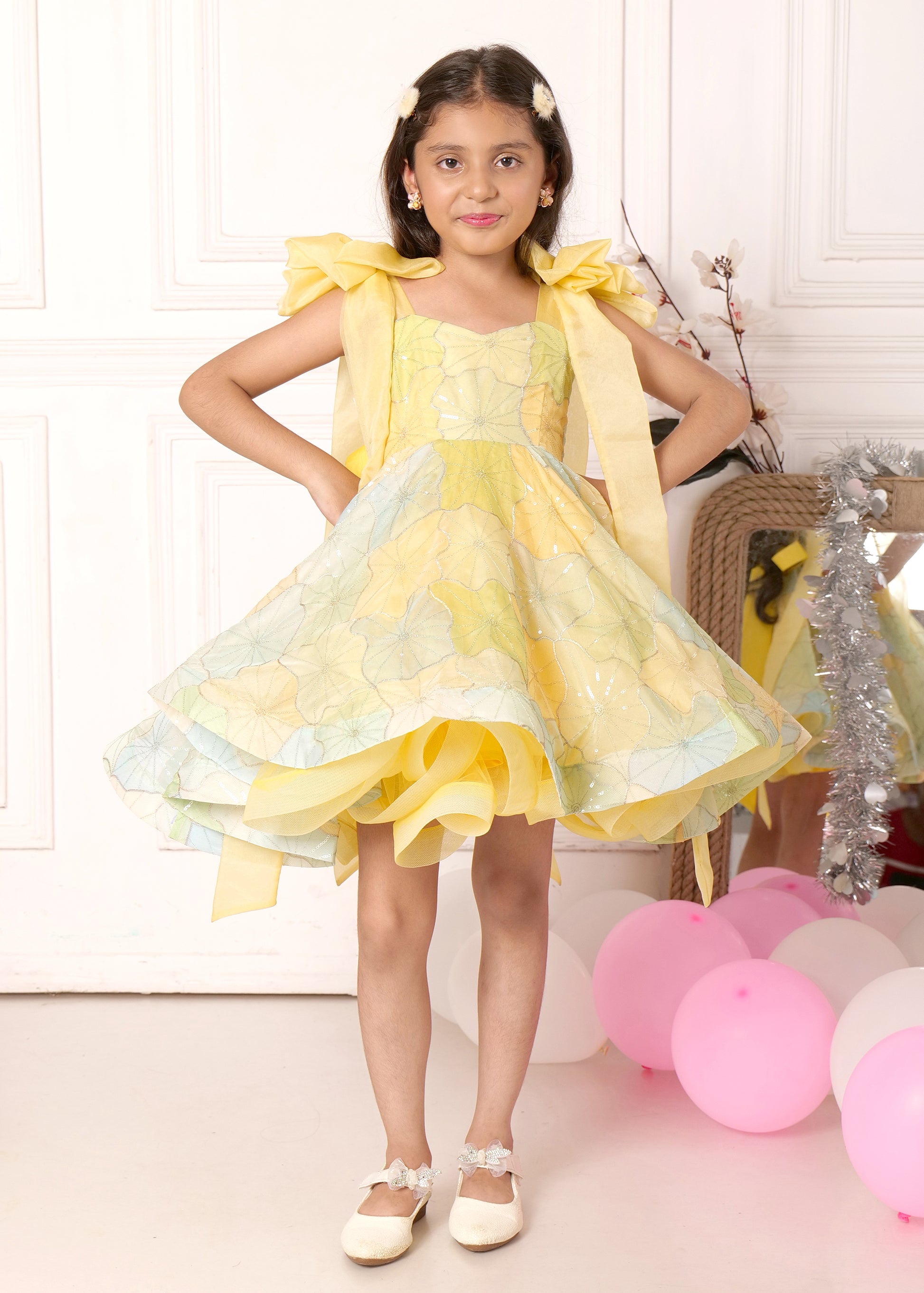 "Adorable girl wearing a pastel yellow organza dress with floral patterns and bow accents, standing confidently in front of pink and white balloons."