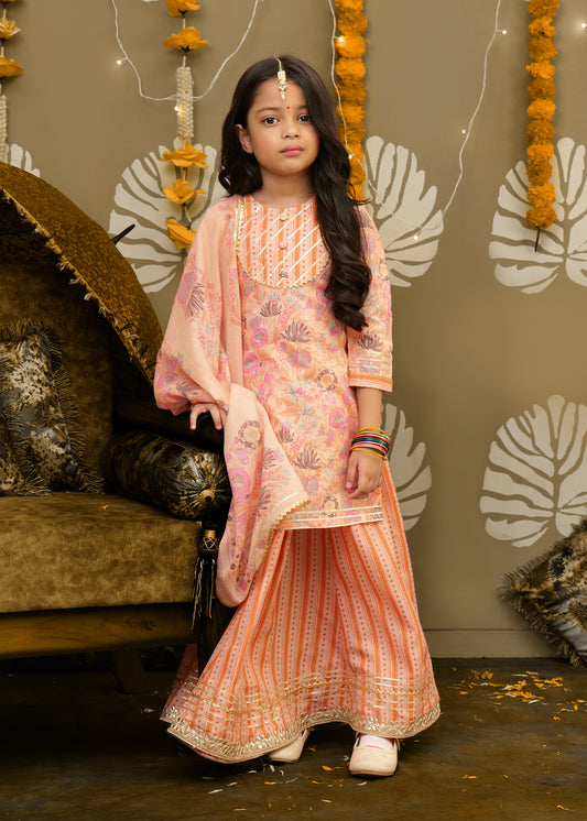 A young girl poses sitting in a peach lehenga with a floral pattern, perfect for festive occasions, styled with bangles and elegant traditional accessories.