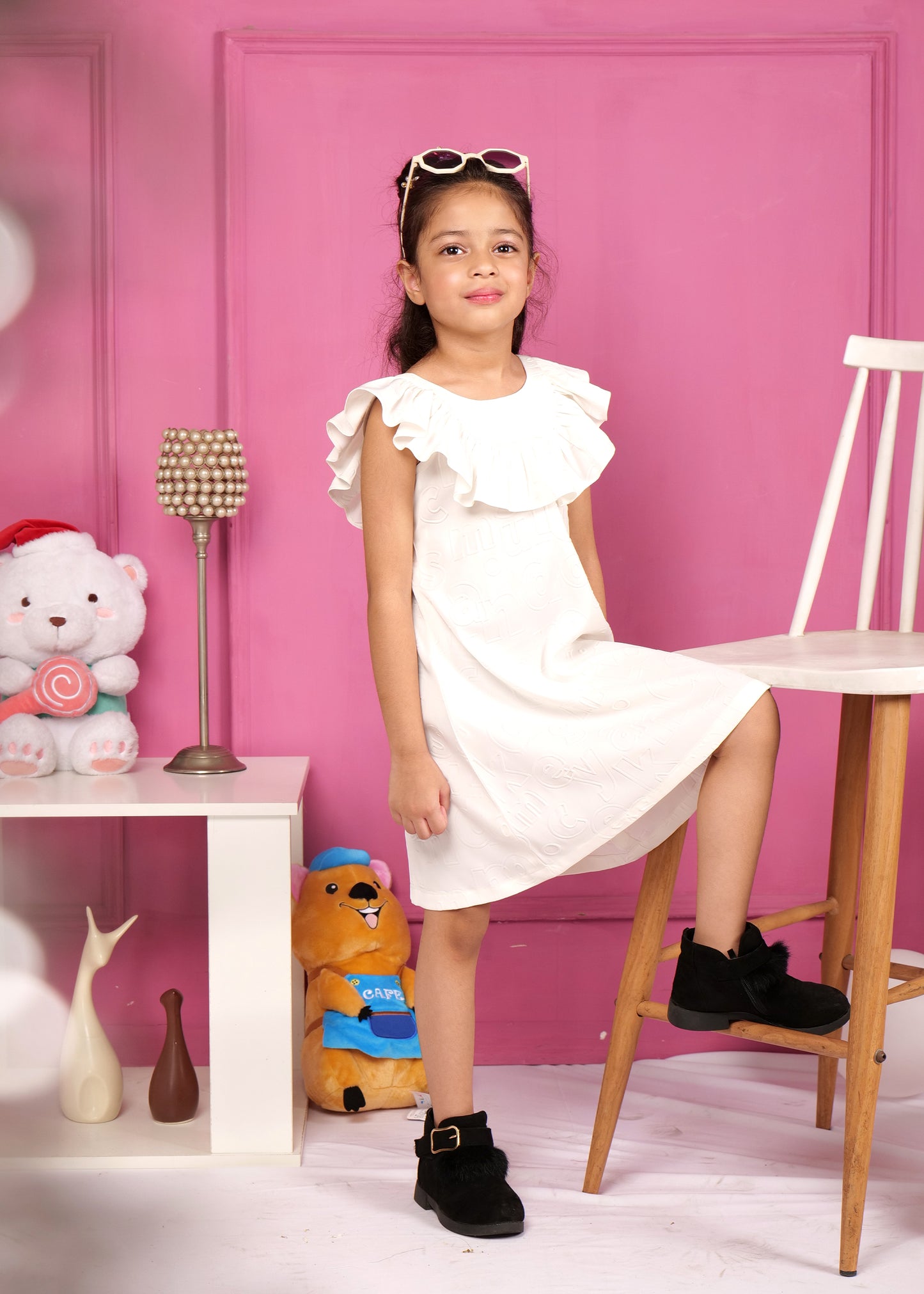 Stylish young girl standing confidently in a white sleeveless dress with ruffled neckline, paired with black boots and sunglasses, posing in front of a pink background.