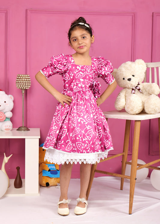 Child model posing in a stylish pink floral print dress with puff sleeves, accessorized with white lace at the bottom, standing in front of a pink backdrop