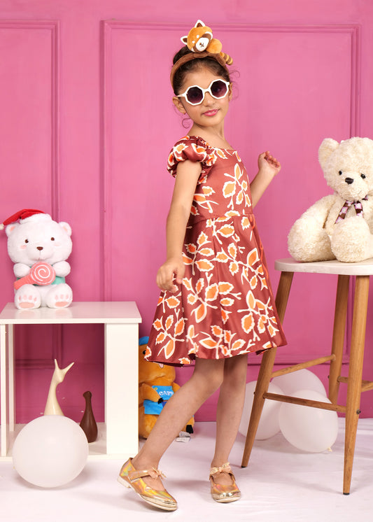 Side view of a girl twirling in a vibrant brown satin dress with orange floral patterns, complemented by a playful headband and golden shoes, set against a pink wall with stuffed animals.