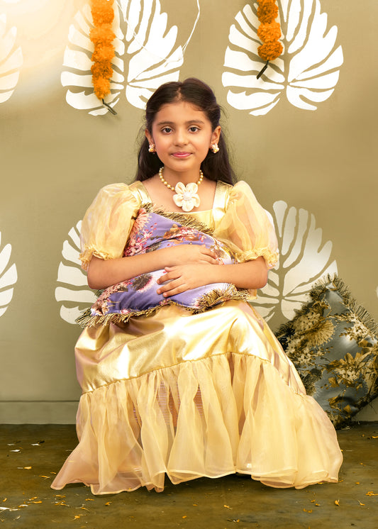 A seated girl in a glamorous golden gown with a puffed skirt and a big bow, holding a floral pillow in a festively decorated room.