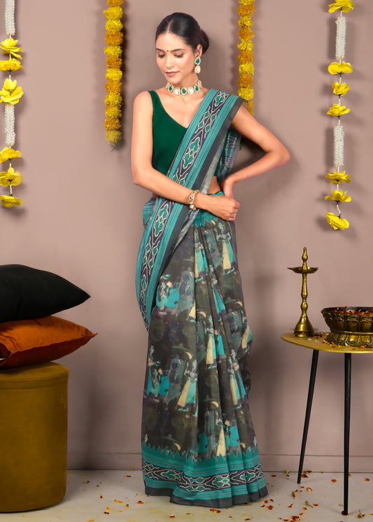 Full view of a model draped in a green saree with black and teal detailing, standing against a traditional backdrop with floral decor