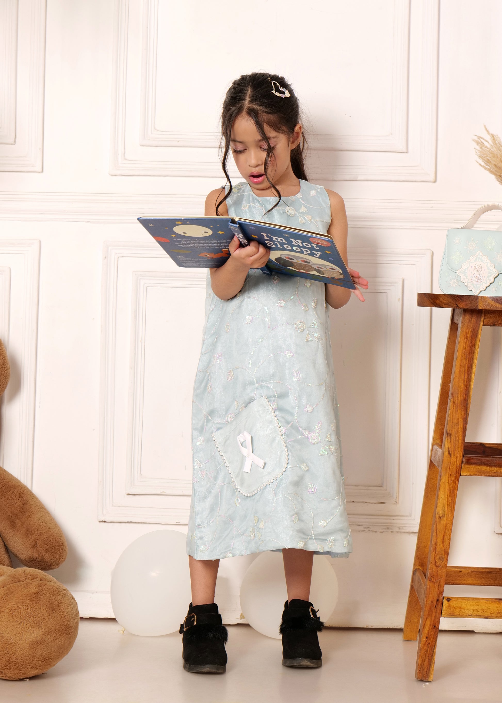 Little girl reading a book while wearing a light blue embroidered dress with a white ribbon pocket detail, paired with black ankle boots, set against a soft indoor background
