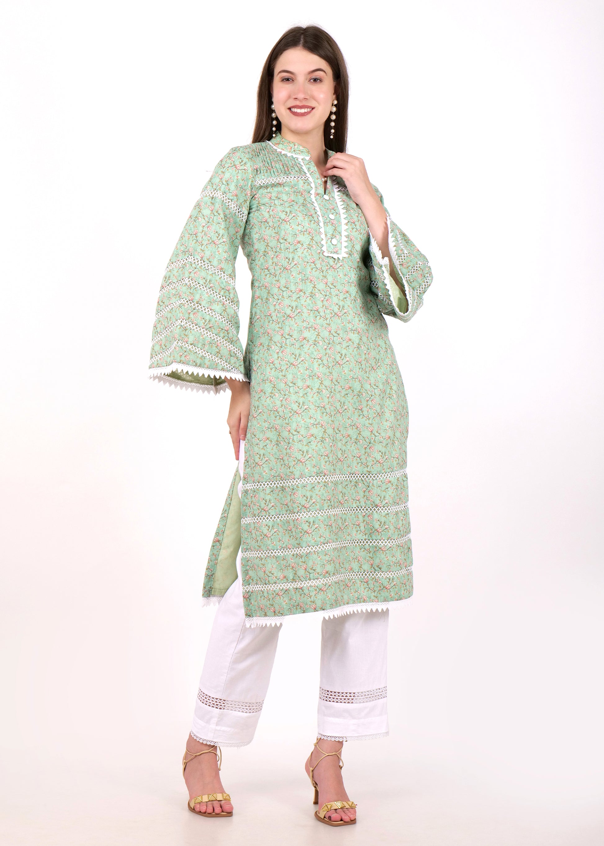  Full view of a woman in a green floral printed kurta with lace borders, paired with white straight pants and heels, smiling gracefully