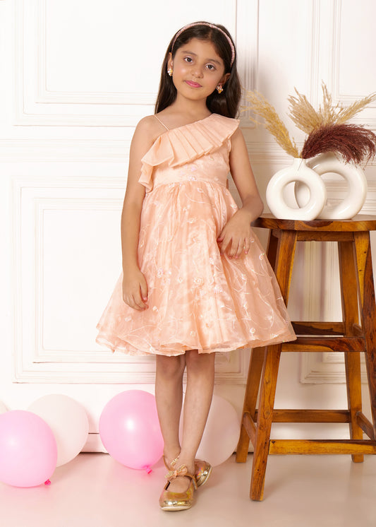 "A girl in a peach one-shoulder dress with floral patterns, standing with one hand on a wooden stool, smiling softly against a neutral backdrop with pink and white balloons on the floor."