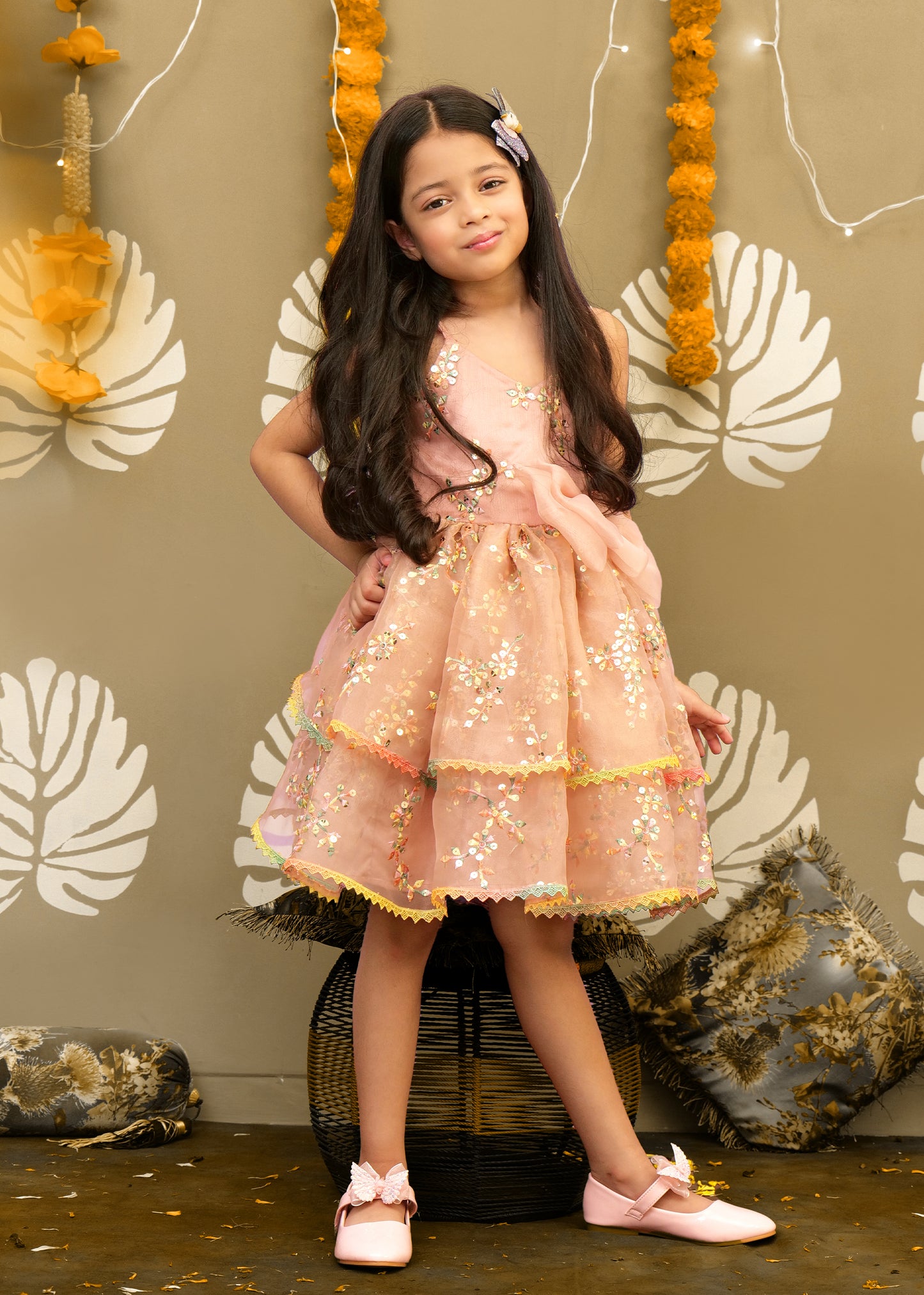 A girl wearing a layered peach floral dress with intricate sequin work, posing confidently in a festive setup with floral decorations.