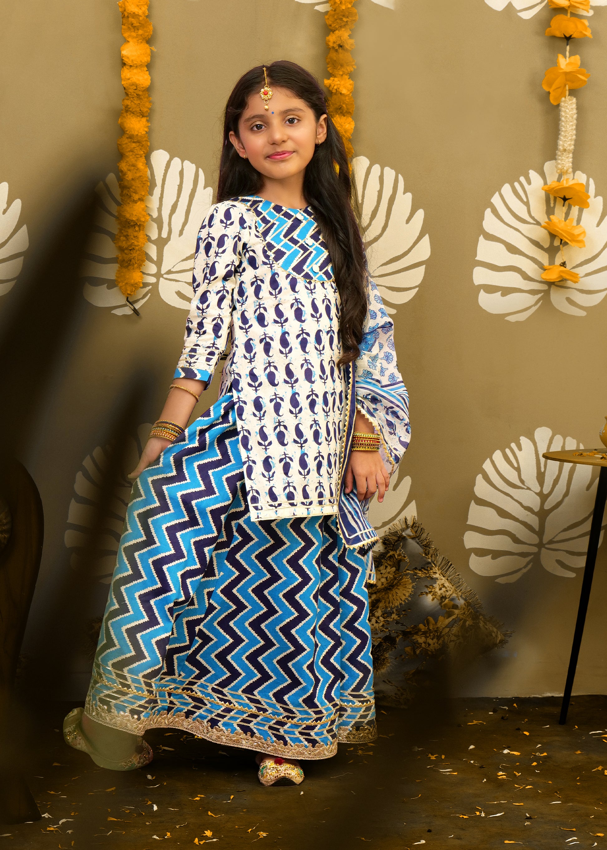 A girl twirling in a blue and white ethnic ensemble with zigzag patterns, paired with a printed palazzo and complementing dupatta, in a traditional setup.