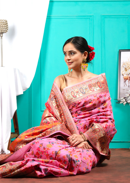 Woman sitting gracefully, wrapped in a pink floral saree with traditional paisley border, adorned with ethnic jewelry